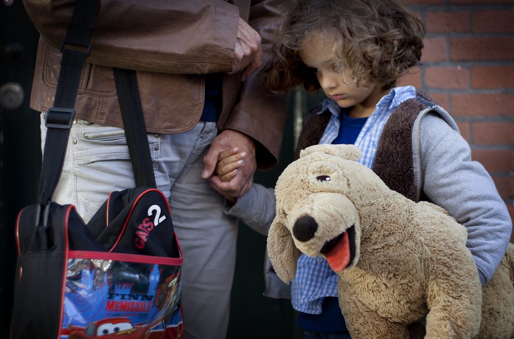 Meisje houdt hand vast van vader met knuffelhond onder haar arm. Vader draagt tas. Meisje kijkt sip.